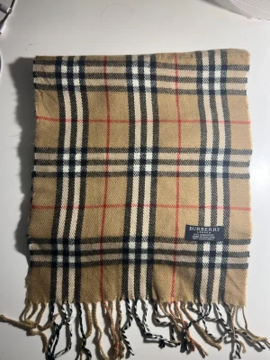 Rutig beige Burberry halsduk i kashmir - Klassisk halsduk från Burberry i 100% kashmir. Beige botten med ikoniskt rutigt mönster i svart, vitt och rött. Tidlös accessoar med fransar i ändarna och vävd Burberry-etikett. Perfekt för dig som vill ha en lyxig och mjuk scarf.