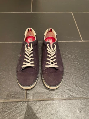 Mörkblå sneakers från Tommy Hilfiger - Snygga mörkblå sneakers från Tommy Hilfiger med vita detaljer och snörning. Skorna har en rund tå, platt sula och är tillverkade i bomullstyg. Insidan är röd med logotyp och hälen har en vit kontrast. Perfekt för dig som gillar klassisk streetstil.