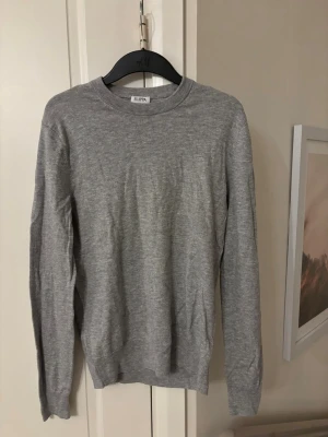 Grå stickad tröja från Filippa K - Snygg grå stickad tröja från Filippa K med rund hals och ribbade muddar vid ärmslut och nederkant. Tröjan har en klassisk, enkel design och är långärmad, perfekt för lager-på-lager under kyliga dagar.