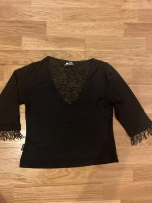 Svart blus med fransar  - Snygg svart blus med djup v-ringning och trekvartsärmar. Ärmsluten har coola fransdetaljer i svart. Gissar på strl Xs eller S. Perfekt nu till nyår!! 