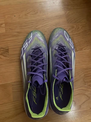 Adidas F50 Elite lila fotbollsskor 43/1/3 - Säljer ett par Adidas F50 fotbollsskor i lila med vita och neongula detaljer. Skorna har snörning och är tillverkade i syntetmaterial för lätt känsla på planen. Perfekta för dig som vill sticka ut med färg och stil på fotbollsplanen. Pris kan diskuteras vid snabb affär.
