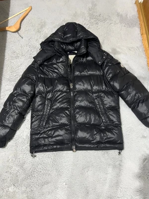 Svart Moncler pufferjacka med huva - Säljer en svart pufferjacka från Moncler i storlek M. Jackan har en stor huva, två sidofickor med dragkedja och coola detaljer som Moncler-logga på ärmen. Materialet är glansigt och känns riktigt lyxigt, perfekt för kalla dagar.