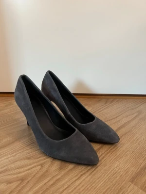 Minellie pumps - Snygga grå pumps från Minelli i storlek 37. Skorna har spetsig tå och är tillverkade i mjuk mocka med en elegant hög klack. Perfekta för dig som gillar stilrena och klassiska skor med en modern touch. Defekt på ena klacken, syns på bild 5. 