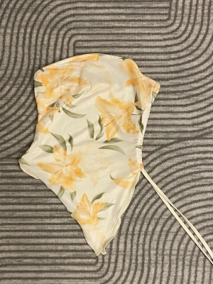 Blommig asymmetrisk tubtopp (SHEIN) - Säljer en snygg tubtopp med asymmetrisk form. Toppen har bandeau-stil och en dekorativ snörning på sidan. Har aldrig använt den för den är för liten:( 