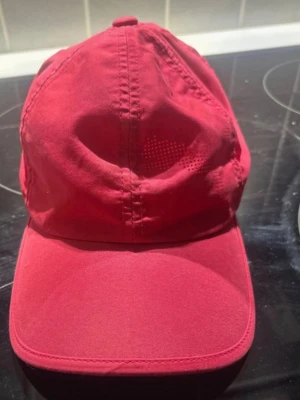 Röd Puma keps med justerbart band - Snygg röd keps från Puma med klassisk böjd skärm och vitt Puma-logo broderat baktill. Kepsen har justerbart band för perfekt passform och är tillverkad i mjukt bomullsmaterial. Perfekt för dig som vill ha en sportig och stilren look.