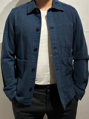 Overshirt J.Lindeberg - Snygg marinblå overshirt från J.Lindeberg i bomullstwill. Jackan har klassisk krage, stora frontfickor och knäppning med svarta knappar. Perfekt att slänga på över en t-shirt för en chill och stilren look. Oanvänd med prislapp kvar. Size Small men passar en Medium.