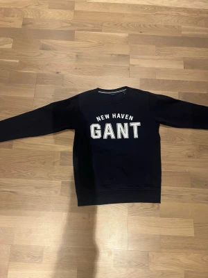 Mörkblå sweatshirt från GANT - Säljer en mörkblå sweatshirt från GANT med vit text 'NEW HAVEN GANT' på bröstet. Tröjan har rund halsringning, långa ärmar och ribbade muddar. Perfekt för dig som gillar klassisk och sportig stil.