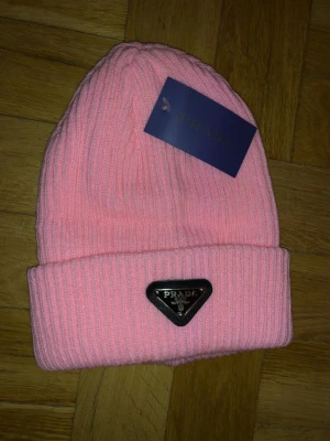 Prada mössa - Säljer en snygg rosa mössa från Prada med ribbstickad design och uppvikt kant. Mössan har en metallbricka med Prada-loggan framtill som ger en lyxig touch. Perfekt accessoar för att lyfta din vinterstil.