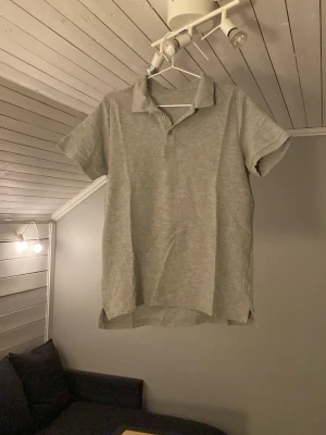 Grå piké t-shirt med krage - Säljer en stilren grå piké t-shirt med klassisk krage och korta ärmar. Tröjan har knappar vid halsen och är gjord i mjukt bomullsmaterial som känns skönt mot huden. Perfekt för dig som gillar enkel och clean stil.