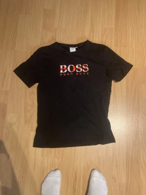 Svart BOSS t-shirt från Hugo Boss - Svart t-shirt från Hugo Boss med stor BOSS-logga i vitt och rött på bröstet. Klassisk rund hals och korta ärmar. T-shirten är gjord i mjuk bomull och har en normal passform som sitter snyggt på kroppen. Perfekt för dig som gillar stilrena märkesplagg.
