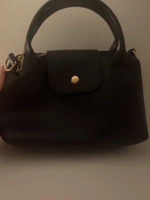 Svart handväska med gulddetaljer - Snygg svart handväska med stilrena gulddetaljer och praktiskt lock med knappstängning. Väskan har två handtag och en klassisk, enkel form som passar till många olika stilar. Materialet är slätt skinn och designen är minimalistisk med fokus på funktion och elegans.