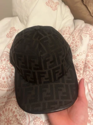 Svart Fendi keps med FF-mönster - Snygg svart keps från Fendi med det klassiska FF-logomönstret över hela kepsen. Justerbar rem baktill med metallspänne där Fendi Roma är ingraverat. Materialet känns lyxigt och har en lätt glans. Perfekt för dig som gillar designeraccessoarer.