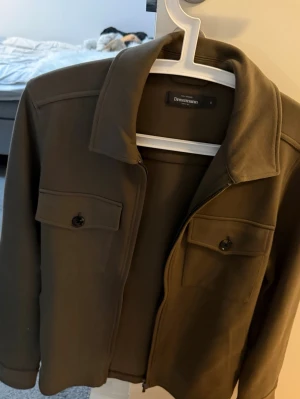 Grön Dressmann overshirt   - Säljer en helt ny olivgrön overshirt från Dressmann i storlek S, med klassisk krage, två bröstfickor med knappar och dragkedja framtill. Jackan har långärmad design och är tillverkad i ett mjukt syntetmaterial som ger en clean och modern look.