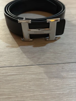 Svart  skärp med H-spänne - Exklusivt svart skärp från  med ikoniskt H-spänne i silverfärgad metall. Skärpet är tillverkat i slätt skinn och har en stilren, klassisk design som lyfter vilken outfit som helst. Perfekt accessoar för dig som gillar lyxiga detaljer.