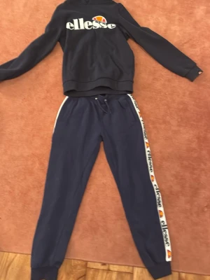 Mörkblå mjukisbyxor från Ellesse - Snygga mörkblå mjukisbyxor från Ellesse med vit logotyp-remsa längs sidorna och snörning i midjan. Byxorna har ribbade muddar vid bensluten och är gjorda i mjukt bomullsmaterial. Perfekta för chill eller träning.