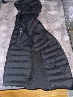 Svart dunväst från Sail Racing - Svart dunväst från Sail Racing med huva och dragkedja framtill. Västen har ett snyggt broderat märke på bröstet och logotyp på framsidan. Quiltad design med paneler och sidopartier i stretchigt material för extra komfort. Perfekt för kyliga dagar.