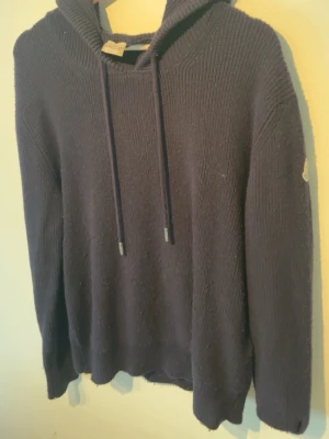 Mörkblå mocler hoodie - Säljer en mörkblå stickad hoodie från Moncler med huva och snörning. Tröjan har långärmade ribbade muddar och Moncler-logga på ärmen. Perfekt för dig som gillar exklusiva märken och vill ha en chill look.