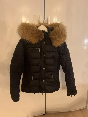 Svart jacka med äkta päls från RockAndBlue - Säljer en svart vinterjacka med stor, fluffig tvättbjörnspäls runt huvan. Jackan har en figurnära passform, dragkedja och knappar framtill samt fickor med dragkedja. Perfekt för kalla dagar när du vill hålla dig varm och snygg.
