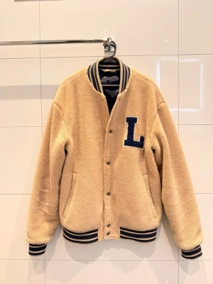 Beige teddyjacka Lexington - Säljer en beige teddyjacka från Lexington i storlek M. Jackan har mörkblå detaljer, randig ribbad krage och muddar, samt en stor blå L på bröstet . Nypris 3000kr säljes för 800kr