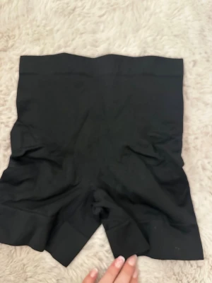 Svarta tighta leggings-shorts - Svarta, korta leggings-shorts med hög midja. De är gjorda i ett stretchigt syntetmaterial som sitter tight mot kroppen och har en enkel, slät design utan synliga detaljer. Perfekta att ha under kläder eller till träning.