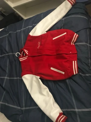 Röd och vit Coca-Cola bomberjacka - Säljer en riktigt snygg röd och vit bomberjacka från Coca-Cola. Jackan har vita ärmar, röd kropp och klassiska ränder på muddar och krage. Broderad Coca-Cola-logga på bröstet och stor logga på ryggen. Två fickor framtill och stängs med tryckknappar.