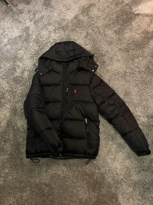 Svart pufferjacka från Polo Ralph Lauren - Svart pufferjacka från Polo Ralph Lauren med huva och dragkedja framtill. Jackan har två sidofickor med dragkedja och en röd Polo-logga på bröstet. Jackan är i storlek S och passar dig som är ca 178. Väldigt fint skick finns inga tecken på andvändning
