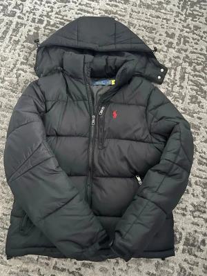 Svart pufferjacka från Polo Ralph Lauren - Svart pufferjacka från Polo Ralph Lauren med huva, dragkedjor och röd broderad logga på bröstet. Jackan har quiltad design och är tillverkad i polyester, perfekt för kalla dagar. Snygg och stilren look med normal passform.
