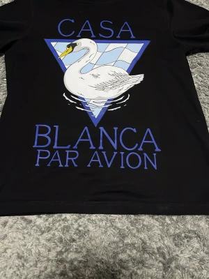 Svart t-shirt Casablanca S - Svart t-shirt från Casablanca med stor grafisk print på framsidan som visar en vit svan och blå text 'Casa Blanca Par Avion'. Triangelbakgrund i blått och vitt. Tillverkad i 100% ekologisk bomull, perfekt för dig som gillar statement-plagg och coola prints.