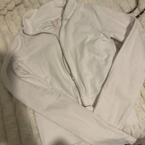 Vit zip-kofta från Nelly - Säljer en vit kofta från Nelly med dragkedja framtill och ribbad krage. Tröjan har lång ärm och är gjord i mjuk bomull, perfekt för lager-på-lager. Enkel och stilren design som passar till många outfits.