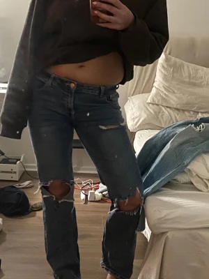 Blå slitna jeans med hål vid knäna - Säljer ett par blå jeans med raka ben och stora slitningar vid båda knäna. Jeansen har klassisk femficksmodell, knappgylf och är midwaist. Materialet är robust denim i bomull och passformen är straight. Perfekta för en avslappnad och trendig look.