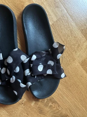 Svarta sandaler med prickigt tyg från Pull&Bear - Svarta sandaler från Pull&Bear med breda band i svart tyg med vita prickar. Sulan är platt och ovandelen har en stor rosett i tyg som ger en cool och lekfull vibe. Perfekt för varma dagar och chill häng.