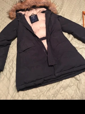 Svart parkasjacka med fuskpäls från Svea - Säljer en svart parkasjacka från Svea i storlek XS. Jackan har beige insida, stor huva med fuskpäls och två stora fickor framtill. Perfekt för kalla dagar med sin varma vaddering och klassiska look. Dragkedja framtill och broderad Svea-logga på bröstet.