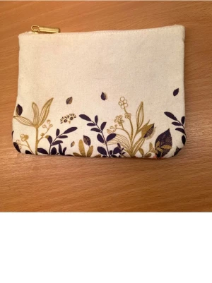 Beige blommig plånbok med dragkedja - Söt liten necessär  i beige bomullstyg med blommigt mönster i mörkblått och guld. Praktisk dragkedja i guld upptill och rymligt fack för småsaker. Perfekt att ha med i väskan för kort, mynt eller smink. Fri frakt 
