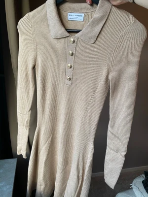 Beige ribbad klänning Holly & Whyte - Snygg beige ribbad stretchig klänning från Holly & Whyte med klassisk krage och fyra guldfärgade knappar framtill. Klänningen har lång ärm och är figurnära upptill med en lätt utsvängd kjol. Perfekt för dig som gillar stilrena och bekväma plagg.