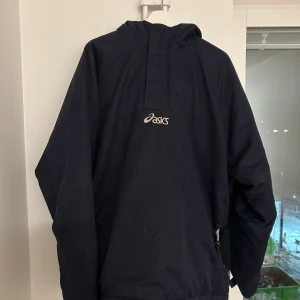 Mörkblå vindjacka från Asics - Säljer en mörkblå vindjacka från Asics med huva och dragkedja i sidan. Jackan har en enkel design med Asics-logga på ryggen och är perfekt för dig som gillar sportigstil och jackan har långa ärmar.