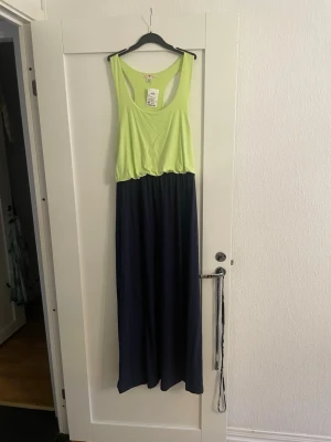 Limegrön och marinblå byxdress - Säljer en snygg byxdress med limegrön topp och marinblå vida byxben. Modellen har djupa ärmhål och resår i midjan för en avslappnad look. Perfekt för dig som vill sticka ut med färg och komfort. Materialet känns mjukt och följsamt.