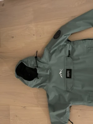 Grön vindjacka från Dope XS - Säljer en grön skidjacka från Dope i storlek XS. Jackan har huva med snörning, stor magficka med lock och Dope-logga framtill samt patch på ärmen. Använt en skidsäsong och är i mycket bra skick!🩷