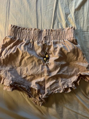 Beige volangshorts från SHEIN - Säljer ett par beige shorts från SHEIN med volangkant och elastisk hög midja. Shortsen har dekorativ snörning med pärlor och tofs framtill. Tillverkade i ett mjukt och luftigt material, perfekta för varma dagar.