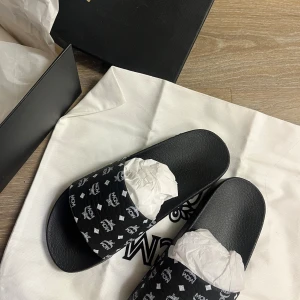 Svarta MCM sandaler - Svarta sandaler från MCM med vit logotyp och mönster över hela remmen. Klassisk slip-on modell med rund tå och platt sula. Tillverkade i syntetmaterial och levereras med originalpåse och låda. Oanvänd 