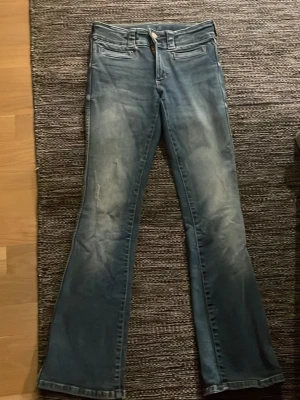 Blå bootcut jeans med dubbla knappar - Snygga blå jeans med bootcut-snitt och dubbla knappar i midjan. Framtill finns två fickor med dekorativa sömmar och baktill två fickor med lock och knapp. Jeansen har en klassisk tvättad look och bälteshällor. Perfekt för dig som gillar retrovibbar. Säljer dessa då de inte är min stil helt. 