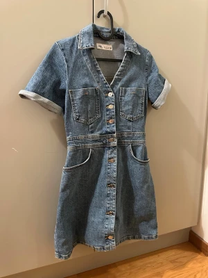 Kort jeansklänning från Zara - Mycket bra skick, endast använd vid 3 korta tillfällen! Säljer en snygg kort jeansklänning från Zara med v-ringning, korta ärmar och knappar framtill. Klänningen har sex fickor samt klassisk skjortkrage. 