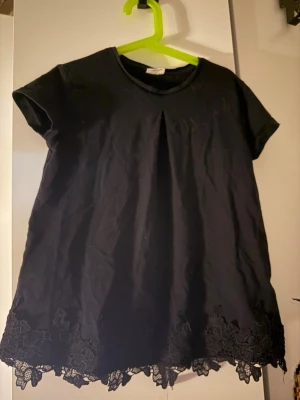 Svart blus med spetskant - Säljer en svart blus med korta ärmar och rund hals. Blusen har en snygg vikt detalj framtill och avslutas med en bred spetskant längst ner som ger en extra touch. Perfekt till jeans eller kjol för en stilren look.