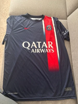 PSG Mbappé matchtröja XL Nike - Säljer en officiell Paris Saint-Germain matchtröja med Mbappé och nummer 7 på ryggen. Tröjan är marinblå med röd och vit rand, PSG-logga och Qatar Airways tryck på bröstet. Materialet är lätt polyester med Dri-Fit teknologi, perfekt för fotboll. Ligue 1 Champion 2023-märke på ärmen.