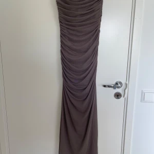 Lång brun draperad klänning med smala axelband - Säljer en långklänning i en mjuk brun nyans med smala axelband och snygg draperad effekt över hela klänningen. Modellen är figurnära och har en diskret dragkedja i sidan. Perfekt för dig som gillar stilrena och eleganta plagg med en twist.