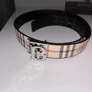 Beige rutig Burberry bälte med B-spänne - Snyggt bälte från Burberry med klassiskt rutigt mönster i beige, brunt och grått. Bältet har ett stilrent metallspänne med Burberrys ikoniska B-logga. Perfekt accessoar för att lyfta din outfit med en lyxig touch.