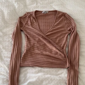 Ribbad omlottopp i beige från Chiquelle - Supersnygg ribbad omlottopp från Chiquelle i en mjuk beige nyans. Toppen har lång ärm, v-ringning och figurnära passform. Perfekt att styla med jeans eller kjol för en trendig look.