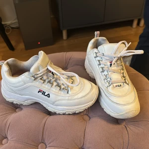 Vita chunky sneakers från Fila, strl 37 - Säljer ett par vita chunky sneakers från Fila i storlek 37. Skorna har en kraftig sula med svarta detaljer undertill, snörning och Fila-logga på sidan och plösen. Ovandelen är i syntetmaterial och mesh för extra komfort.