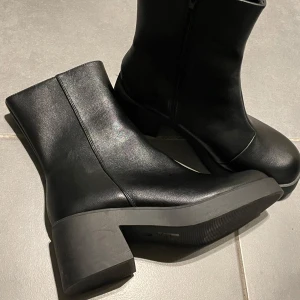 Svarta boots med klack och dragkedja - Säljer ett par svarta boots i skinnimitation med chunky klack och rund tå. Skorna har dragkedja på insidan för enkel påtagning och en stilren, minimalistisk look. De är helt oanvända, endast prövade.