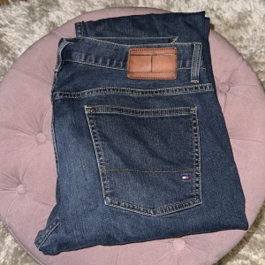 Tommy Hilfiger blå jeans 36/36 - Mörkblå jeans från Tommy Hilfiger i klassisk femficksmodell. Snygga kontrastsömmar och diskret logga på bakfickan. Jeansen har normal passform och är tillverkade i slitstarkt denimtyg av bomull. Perfekta för dig som gillar stilrena och tidlösa jeans.