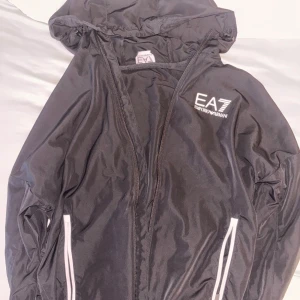 Svart vindjacka EA7 Emporio Armani - Svart vindjacka från EA7 Emporio Armani med huva och vita detaljer vid fickorna. Jackan har dragkedja framtill och logga på bröstet. Perfekt för dig som vill ha en stilren och sportig look.